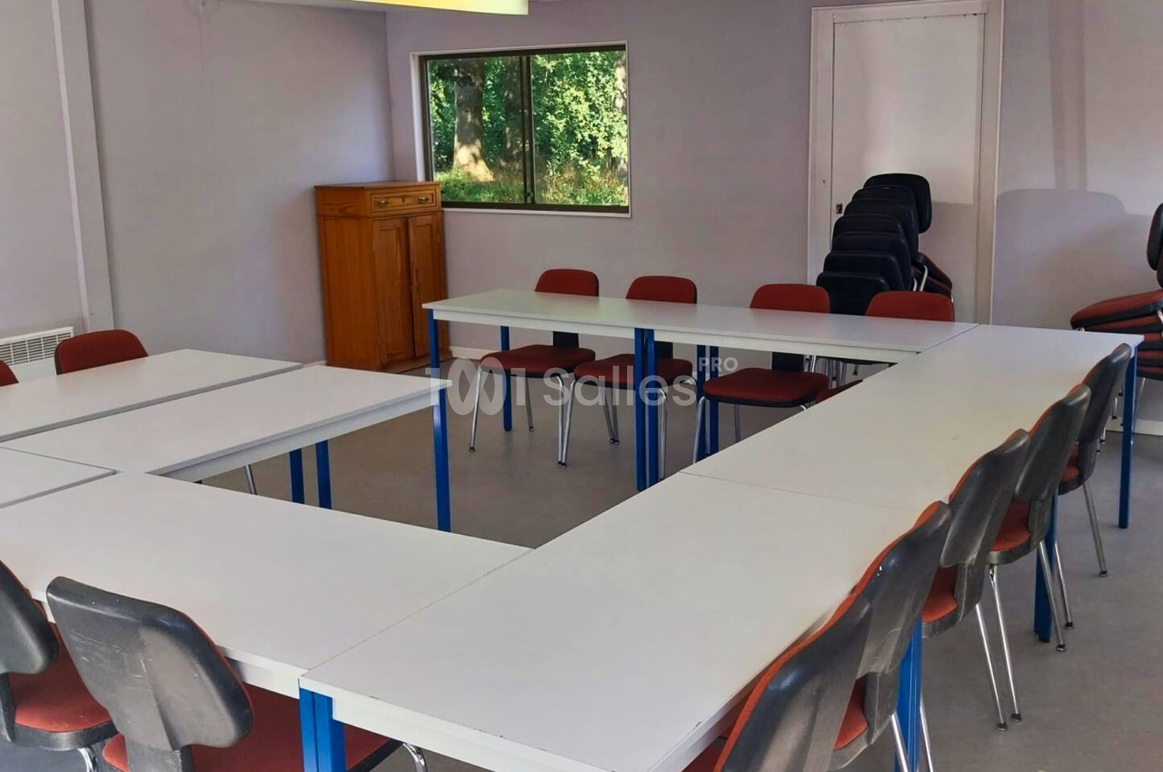Salle de réunion lumineuse avec des tables disposées en U, entourées de chaises rouges et vue sur un espace extérieur.