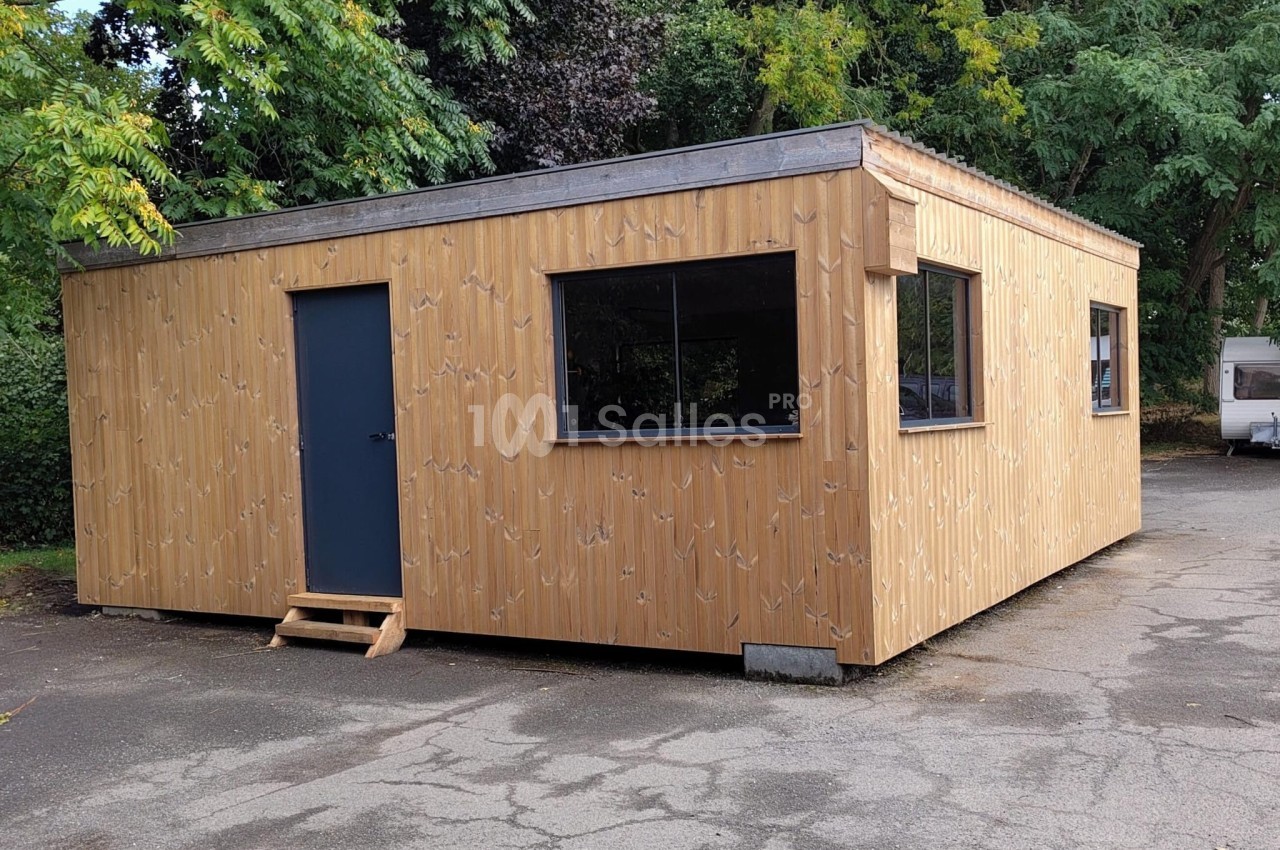 Un bâtiment en bois avec une porte et des fenêtres, situé sur un sol asphalté entouré de végétation.