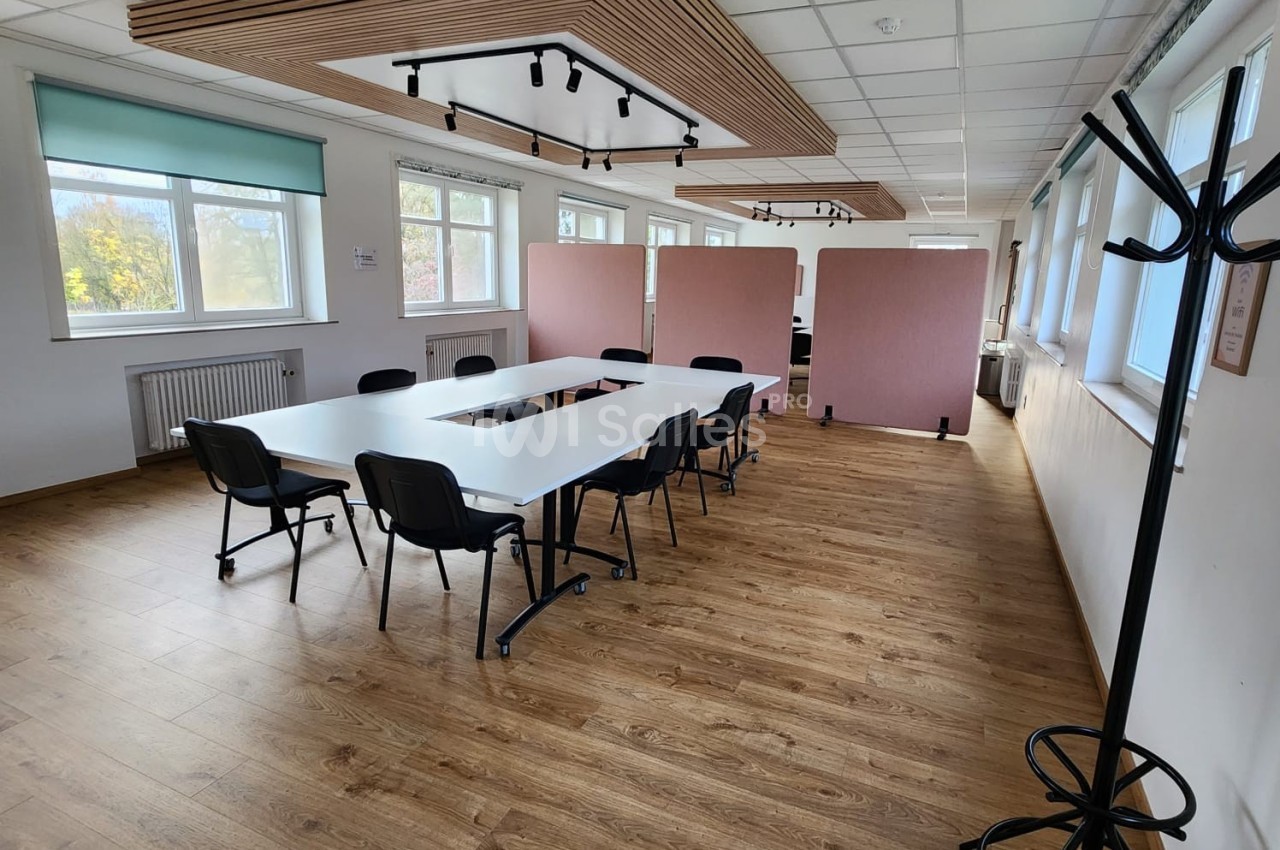 Salle de réunion lumineuse avec tables blanches, chaises noires, parquet en bois et cloisons roses en arrière-plan.