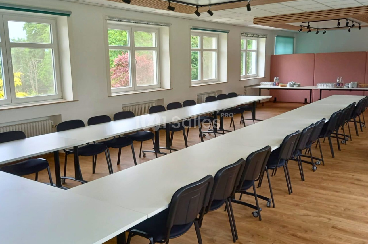Salle de réunion lumineuse avec des tables disposées en U, des chaises noires et un sol en bois clair.