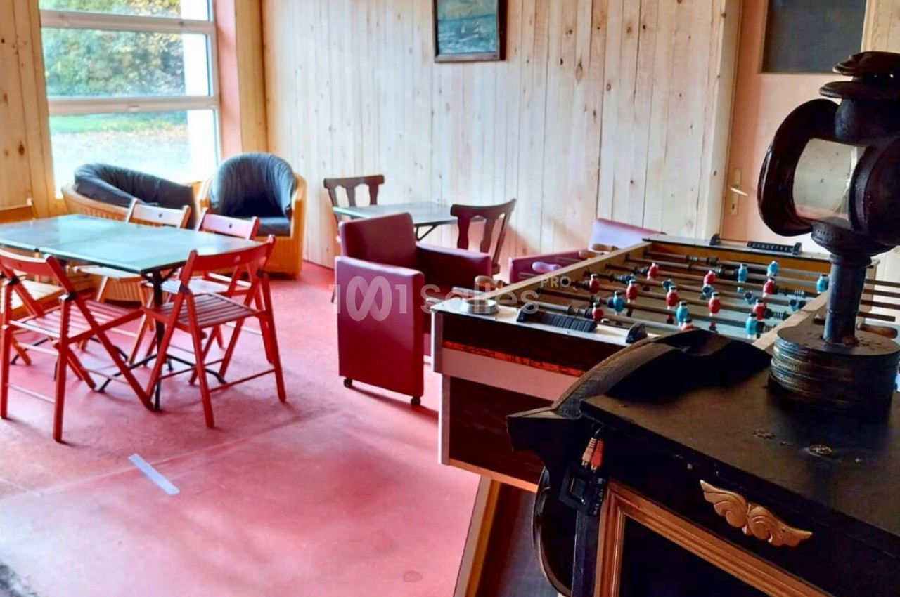 Salle de détente avec baby-foot, tables en bois, chaises pliantes et fauteuils, éclairée par une grande fenêtre.