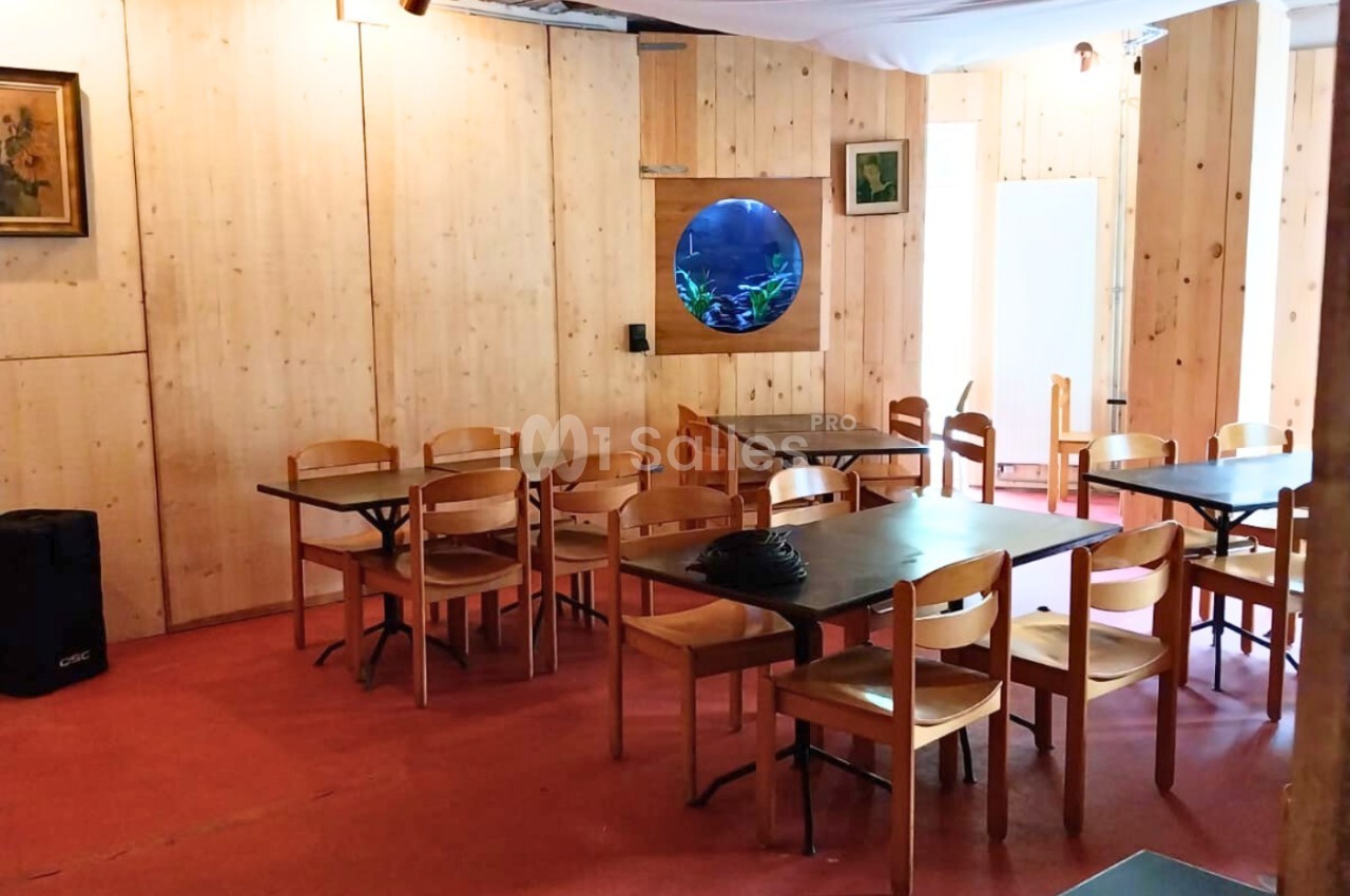 Salle avec des murs en bois, des tables et chaises en bois clair, un aquarium rond et un plafond avec poutres apparentes.