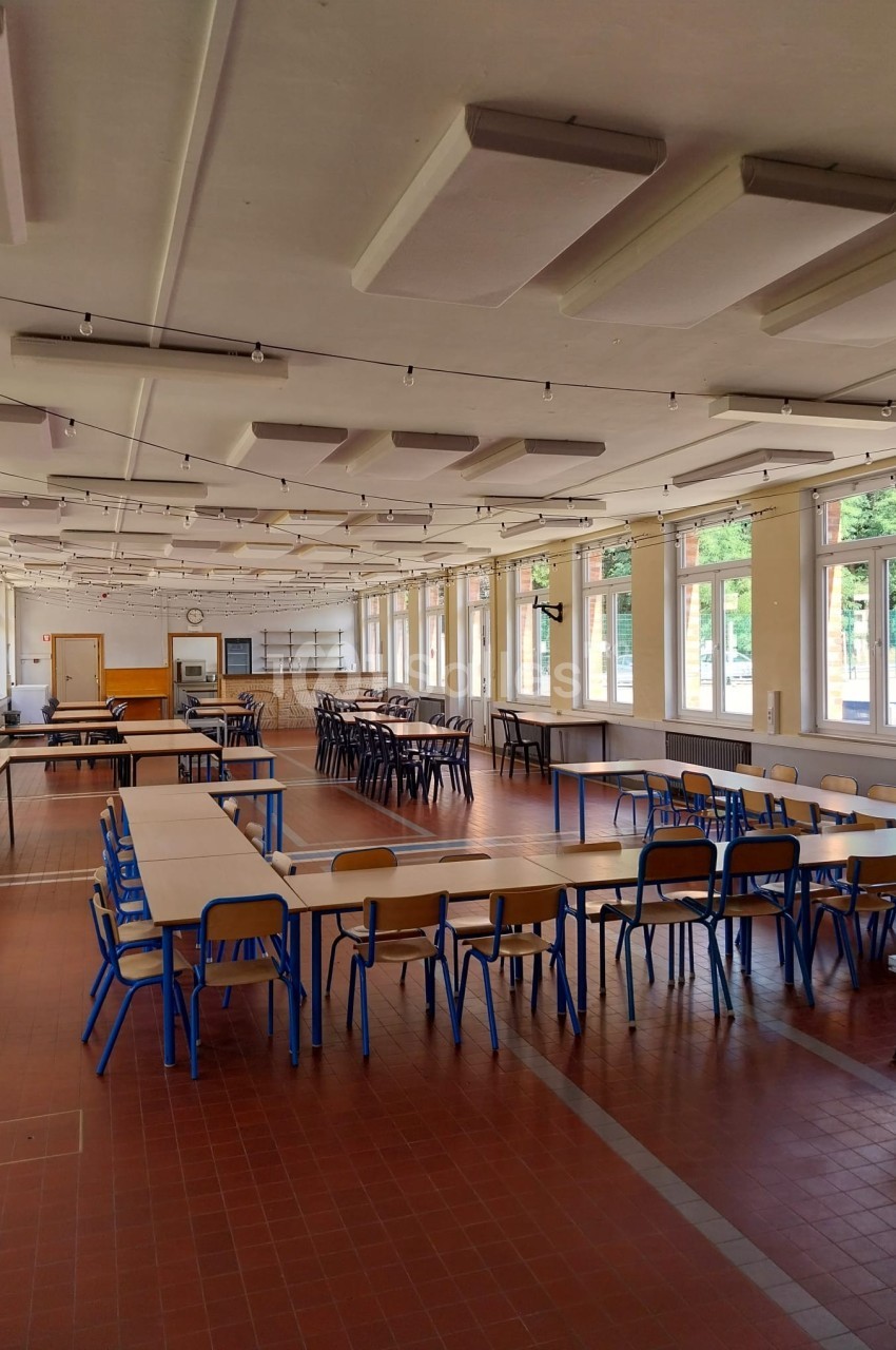 Salle de cantine lumineuse avec de grandes fenêtres, des tables alignées et des chaises bleues sur un sol en carrelage rouge.