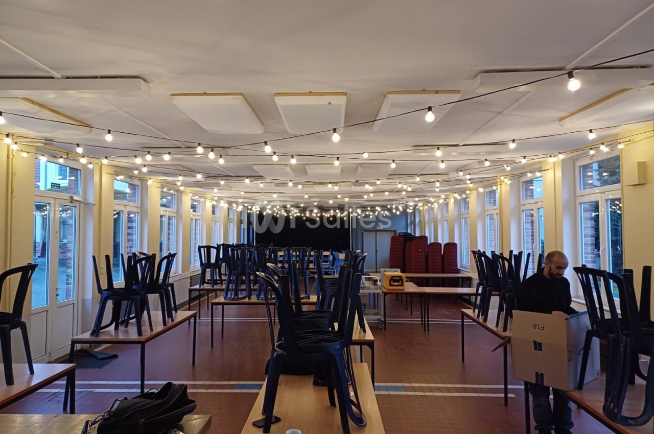 Salle lumineuse en préparation, avec des chaises empilées sur des tables et une personne portant un carton.