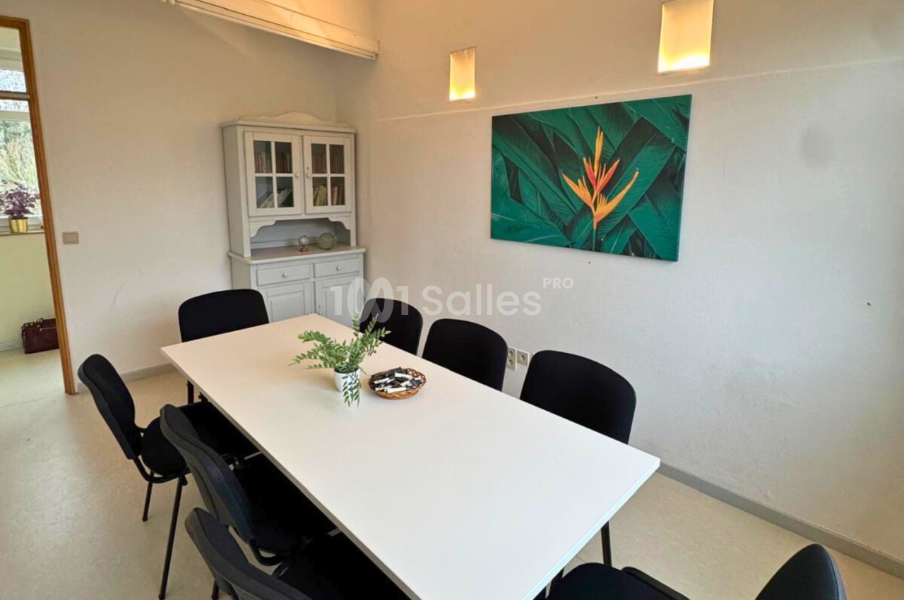 Salle de réunion lumineuse avec une table blanche, des chaises noires, un buffet et un tableau représentant une fleur…