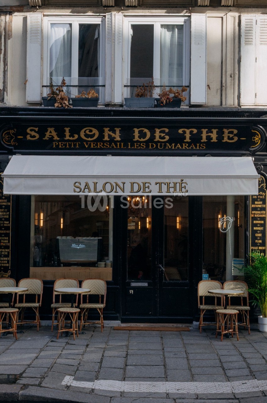 Façade d'un salon de thé parisien avec une devanture noire, une enseigne beige et des chaises en terrasse.