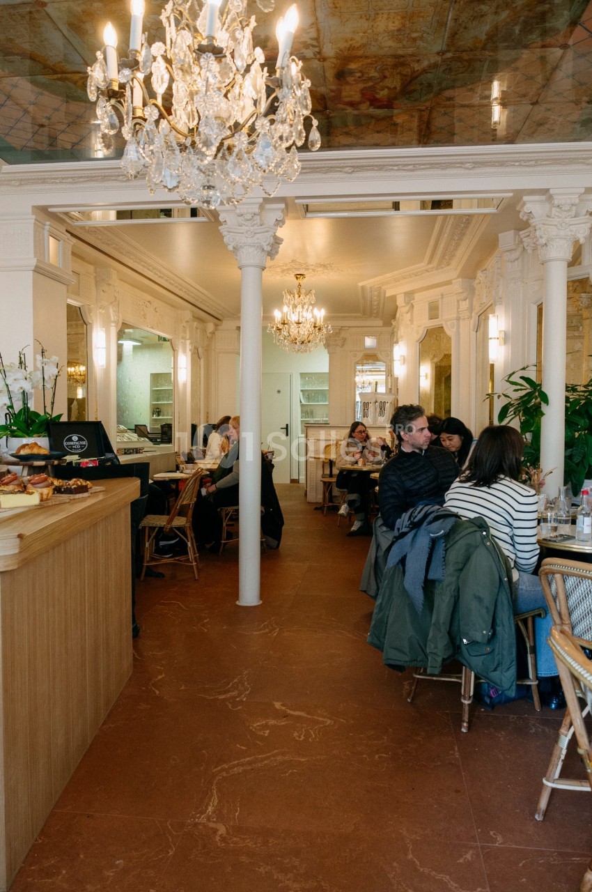 Intérieur d'un café lumineux avec des clients assis, un comptoir à pâtisseries et des lustres décoratifs.