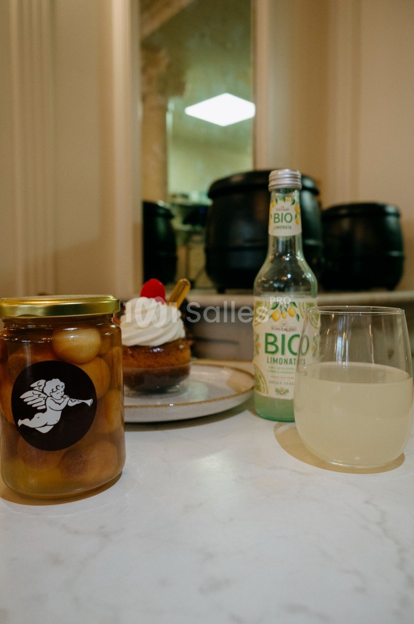 Un pot de fruits au sirop, une tartelette, une bouteille de limonade bio et un verre sur une table en intérieur.