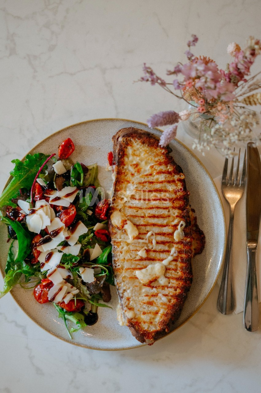 Assiette avec une tranche de pain grillé au fromage et une salade verte garnie de tomates cerises et de sauce blanche.