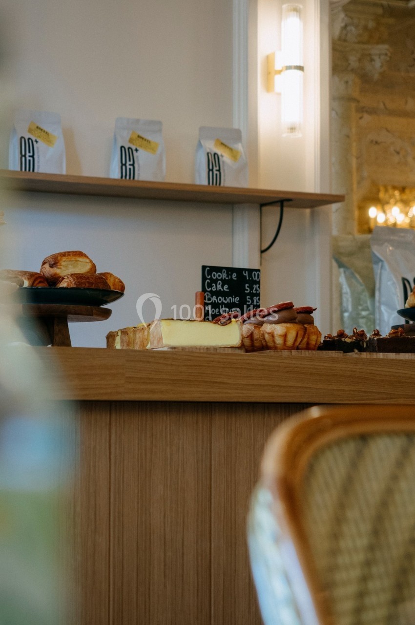 Comptoir de boulangerie avec pâtisseries, pain et ardoise indiquant les prix, éclairage chaleureux en arrière-plan.