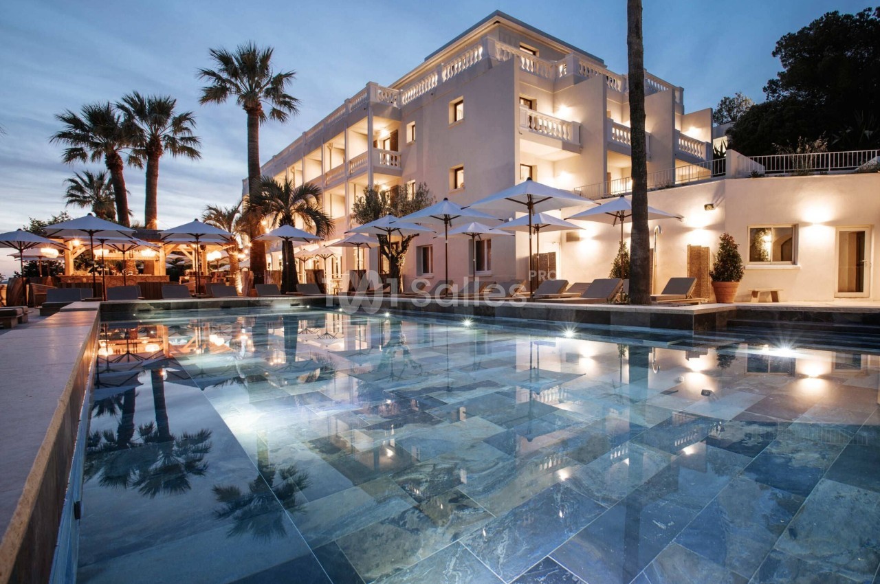 Piscine éclairée au crépuscule devant un bâtiment élégant entouré de palmiers et de parasols.