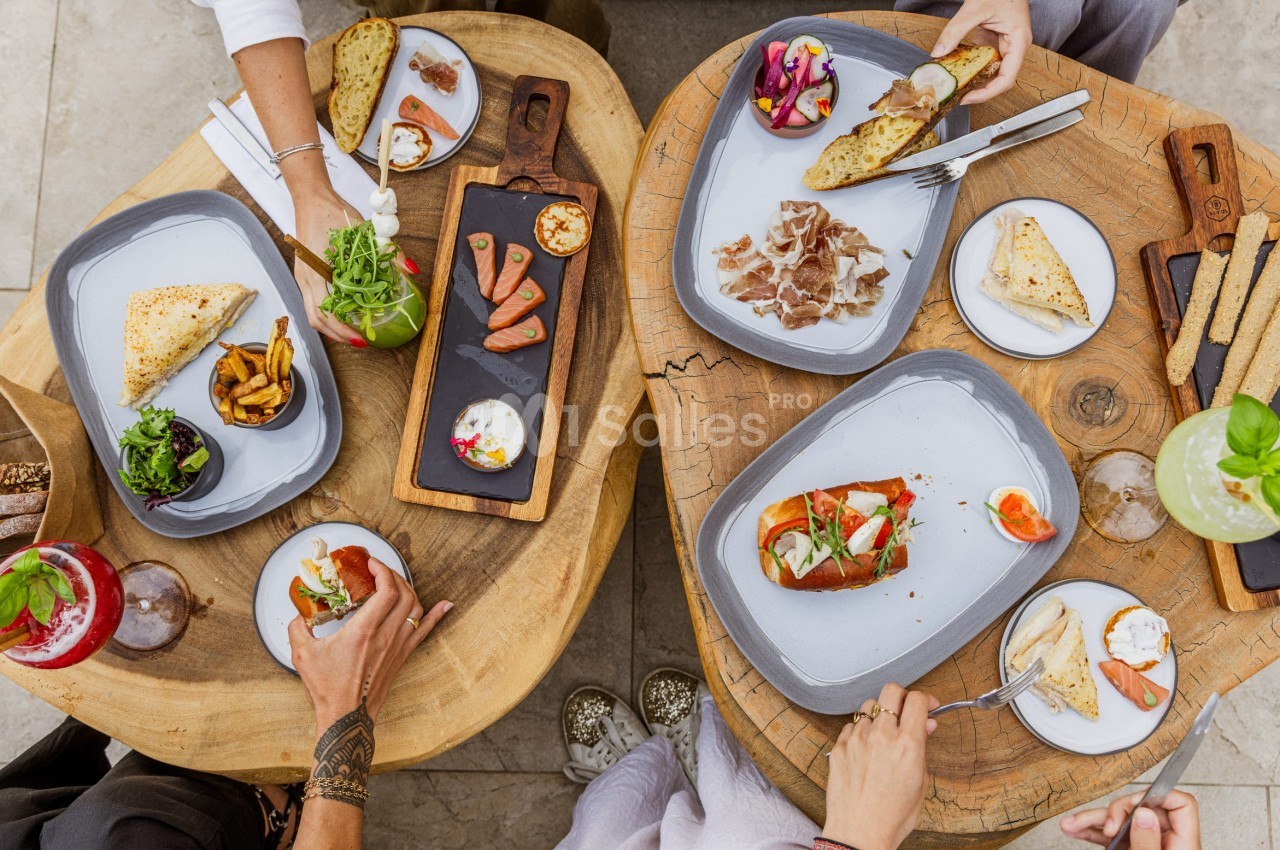 Vue de dessus de deux tables en bois avec des assiettes de tapas, boissons et mains en train de partager un repas.