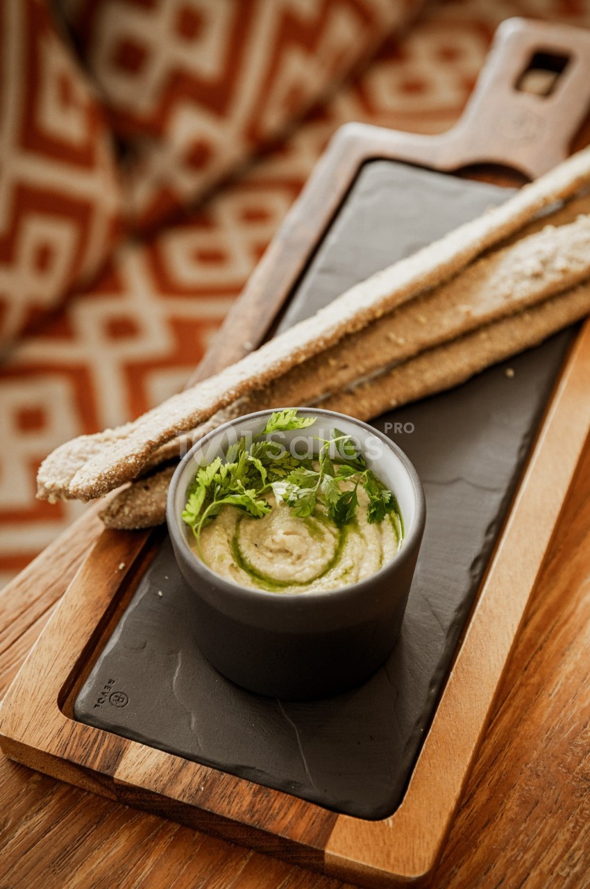 Bol de houmous garni d'herbes fraîches, accompagné de gressins sur une planche en bois.