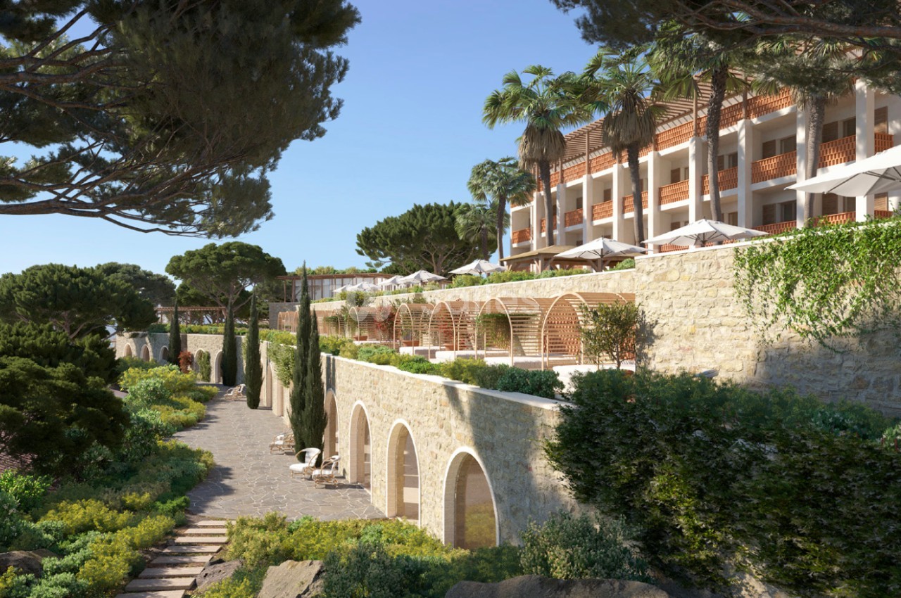 Chemin bordé de végétation menant à un bâtiment en pierre avec terrasses et parasols, entouré de pins et palmiers.