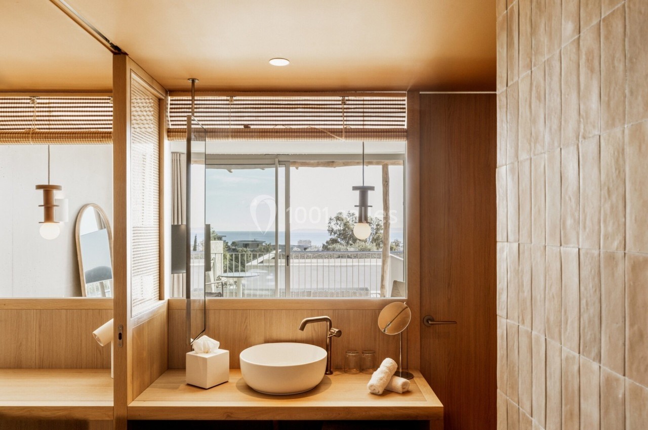 Salle de bain lumineuse avec lavabo rond, miroir ovale, et vue sur l'extérieur à travers une grande fenêtre.
