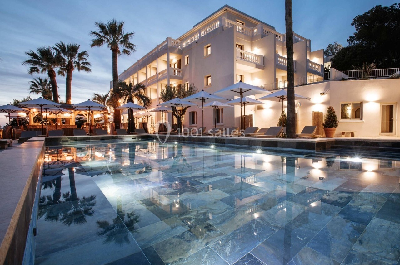 Piscine éclairée au crépuscule devant un bâtiment élégant entouré de palmiers et de parasols.
