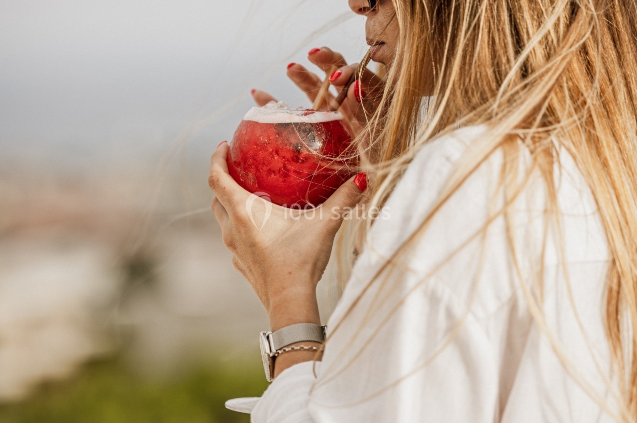 Femme buvant un cocktail rouge avec une paille, en extérieur, cheveux blonds au vent.