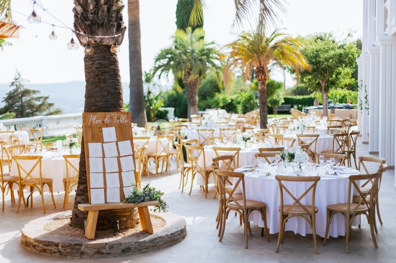 Tables de mariage dressées en extérieur avec des chaises en bois, un plan de table appuyé contre un palmier.