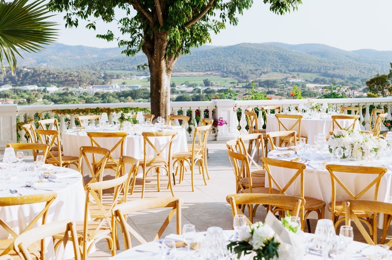 Tables dressées pour un événement en plein air, avec chaises en bois, nappes blanches et vue sur des collines verdoyantes.