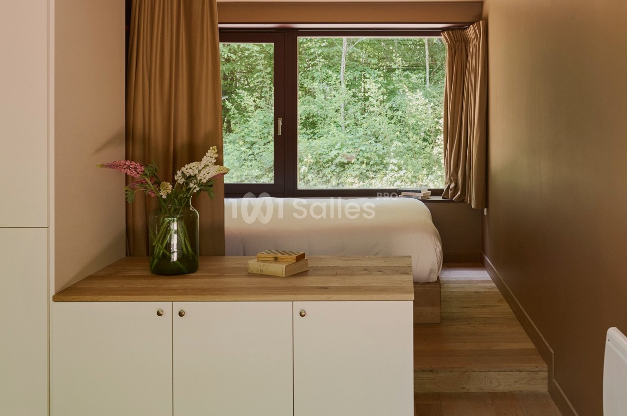Chambre lumineuse avec lit, grandes fenêtres donnant sur une forêt, meuble en bois clair et vase de fleurs.