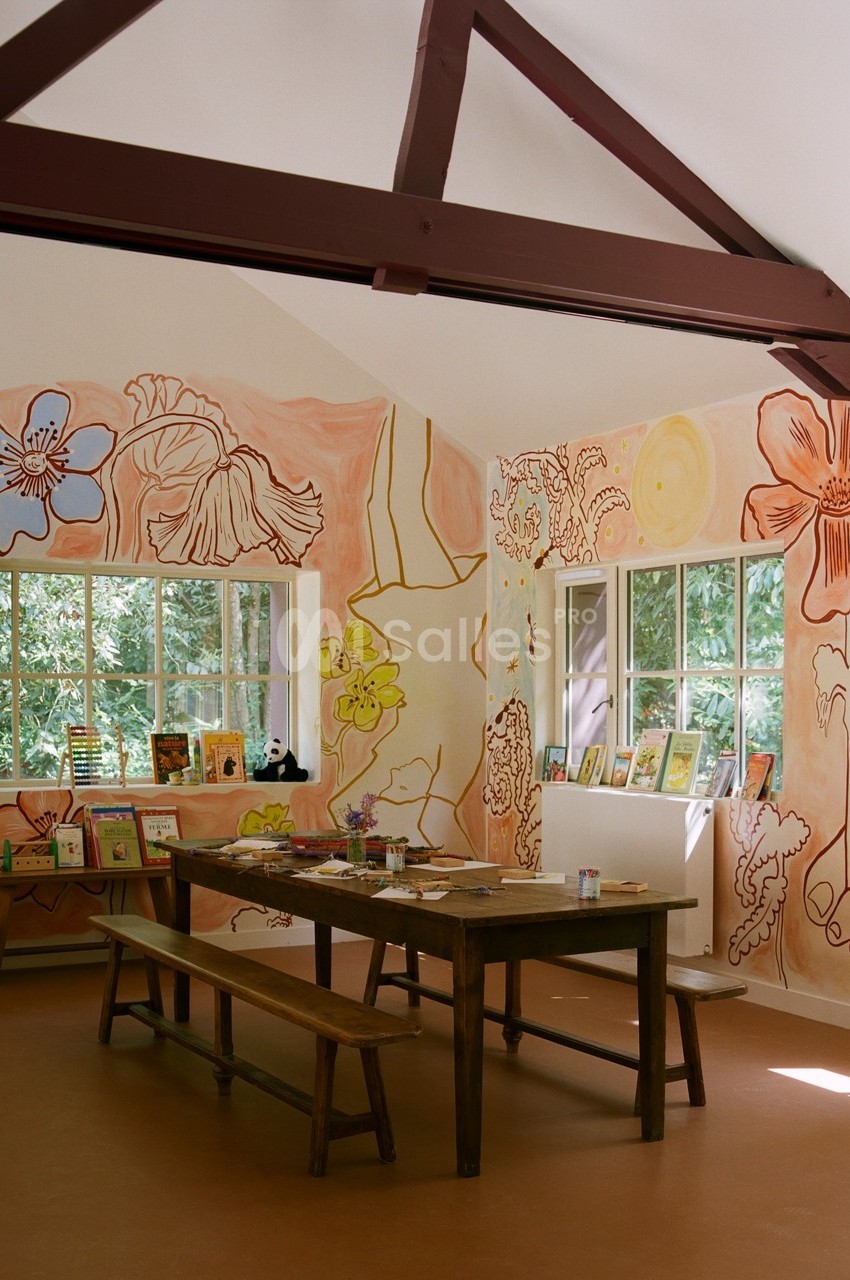 Salle lumineuse avec des murs décorés de peintures florales, tables en bois et fenêtres donnant sur un espace verdoyant.