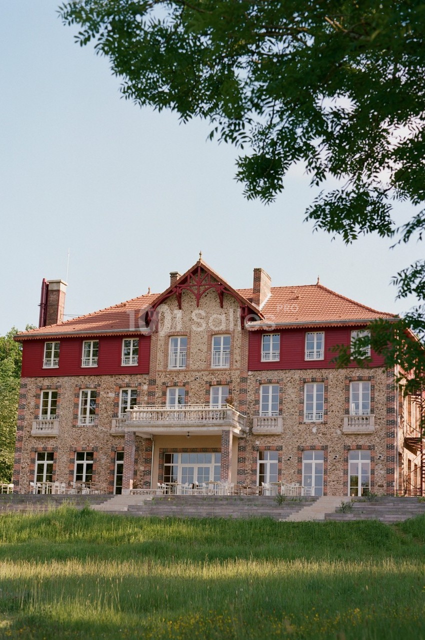 Façade d'un grand bâtiment en briques rouges et pierres, entouré de verdure sous un ciel dégagé.