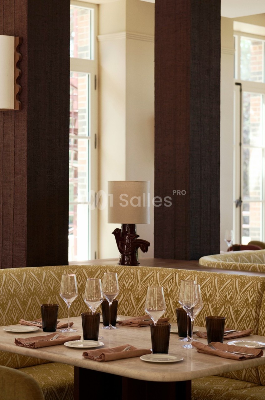 Banquette beige avec table dressée, verres et serviettes, dans un restaurant lumineux avec grandes fenêtres.