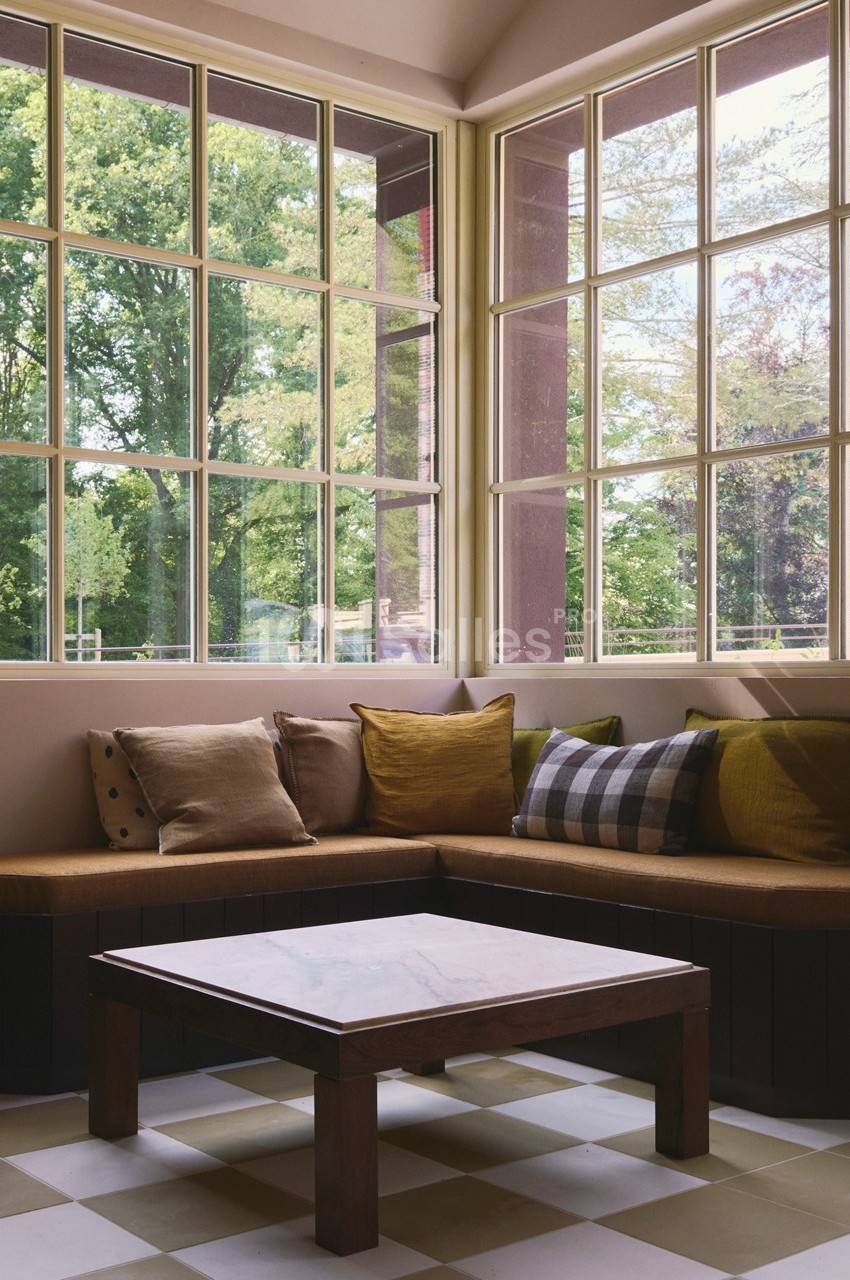 Coin salon lumineux avec banquette en bois, coussins colorés et table basse, donnant sur un jardin verdoyant.