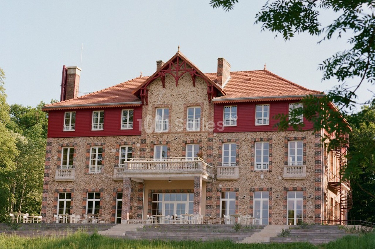 Façade d'un grand bâtiment en briques avec des détails rouges, entouré de verdure et éclairé par la lumière naturelle.