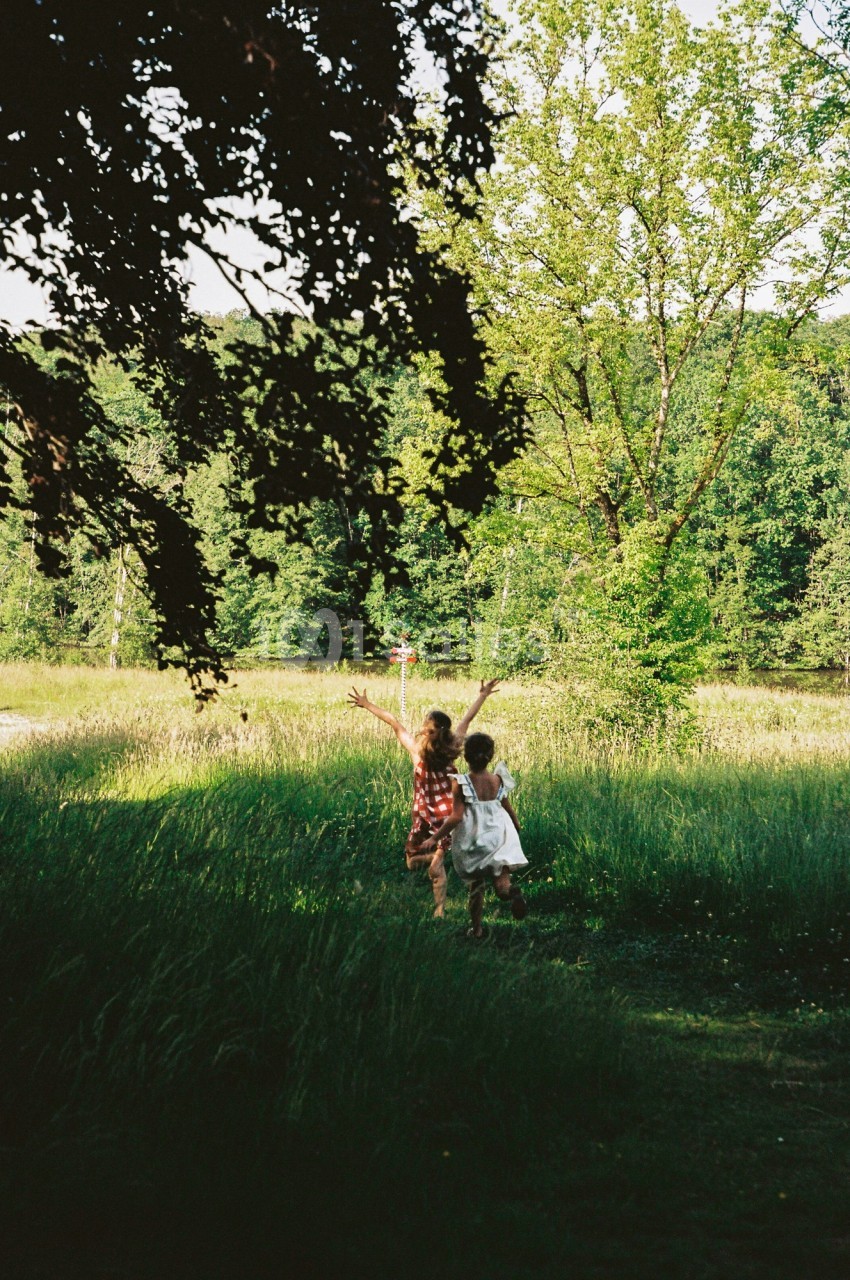 Deux enfants courent dans une prairie verdoyante, entourés d'arbres et baignés par une lumière naturelle.
