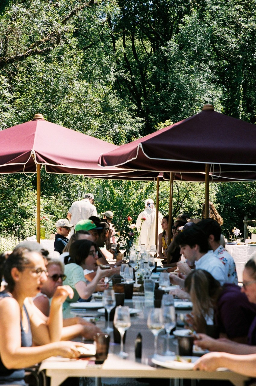 Des personnes déjeunent à de longues tables sous des parasols rouges dans un cadre verdoyant.