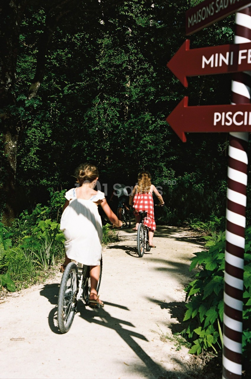 Deux enfants à vélo sur un chemin forestier, près de panneaux indiquant des directions touristiques.