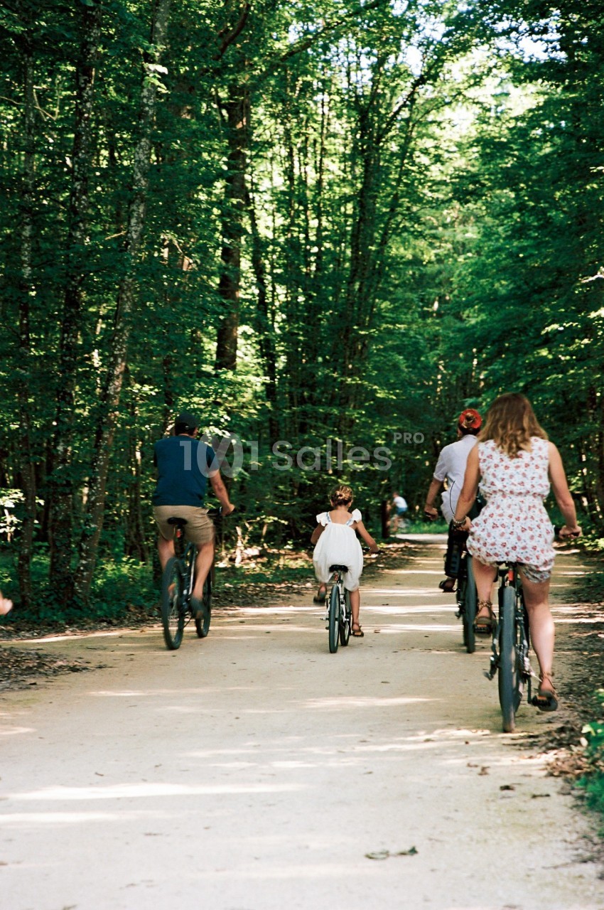 Des personnes, dont une enfant, font du vélo sur un chemin forestier ombragé par une journée ensoleillée.