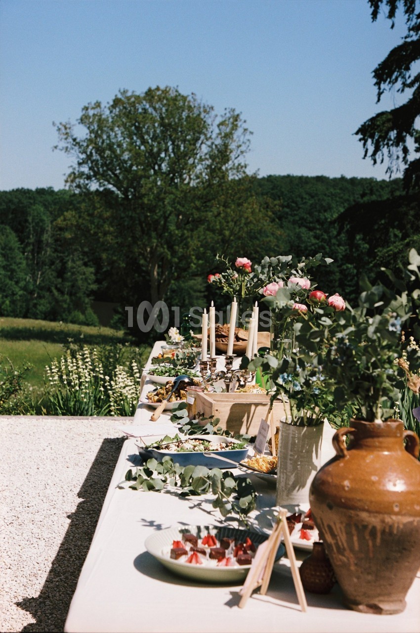 Table dressée en extérieur avec plats variés, bougies et décorations florales, entourée de verdure.