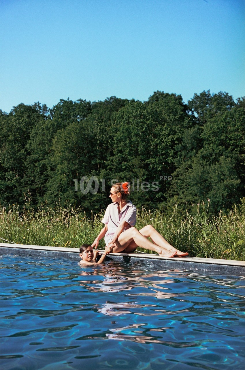 Une femme assise au bord d'une piscine aide un enfant dans l'eau, avec une forêt en arrière-plan.