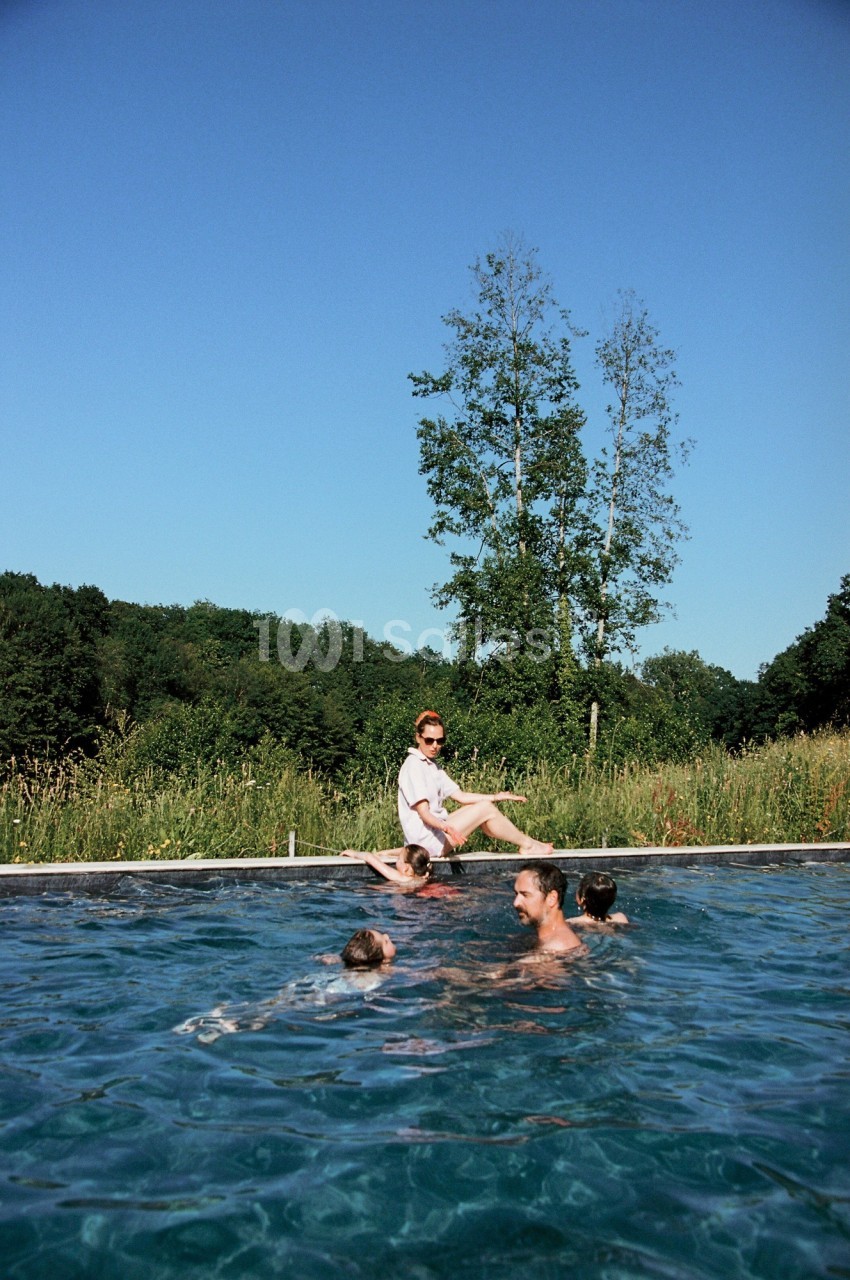 Des personnes se baignent dans une piscine extérieure entourée de verdure sous un ciel bleu.