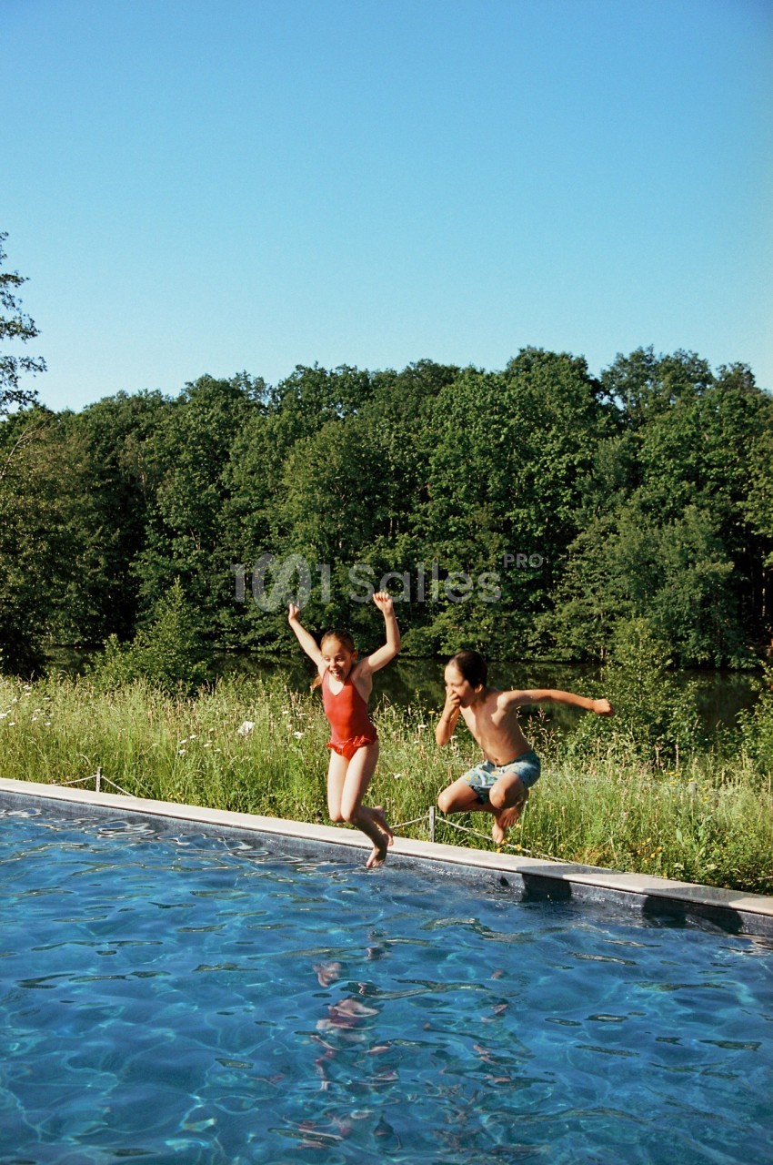 Deux personnes sautent dans une piscine en plein air, entourée de verdure et d'arbres sous un ciel dégagé.