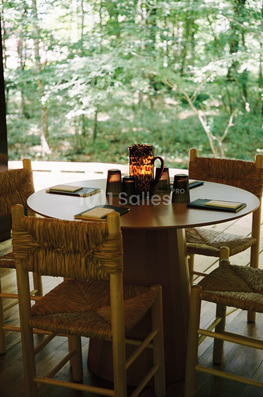 Table ronde en bois entourée de chaises en paille, avec des carnets et une carafe au centre, vue sur une forêt.