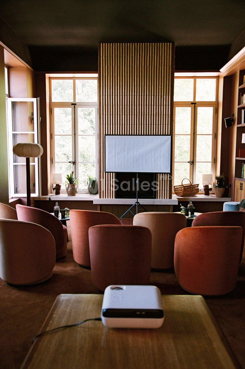 Salle aménagée avec des fauteuils colorés, un projecteur et un écran blanc devant de grandes fenêtres.