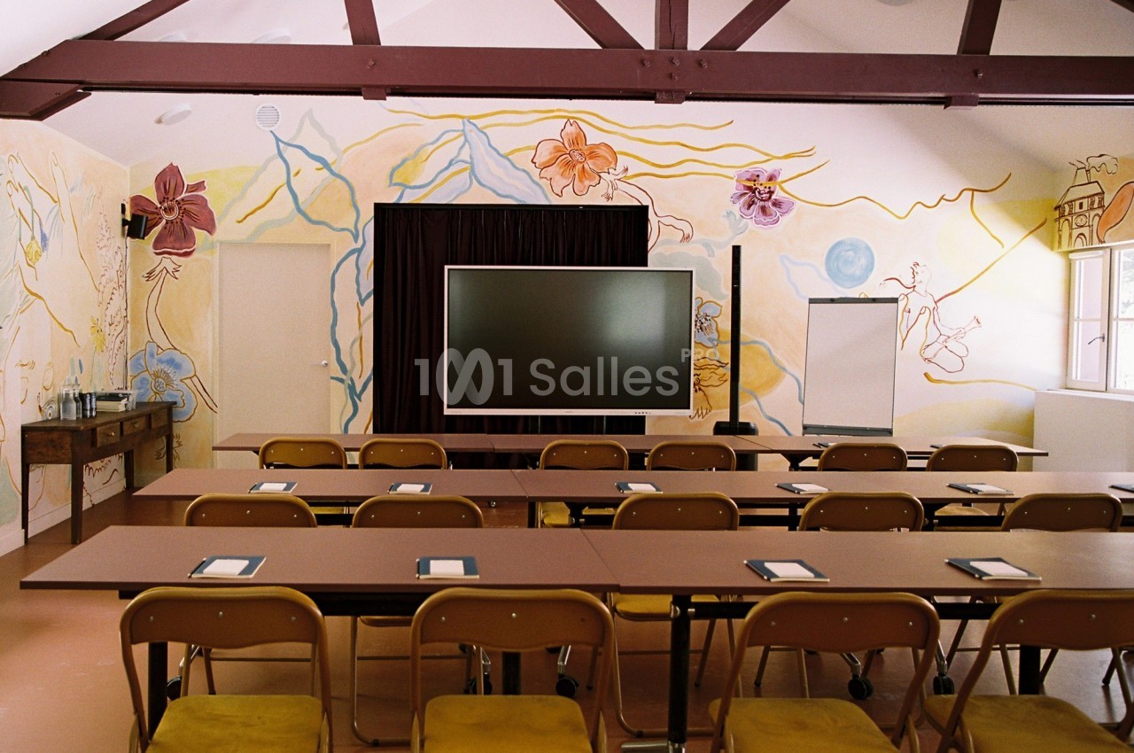 Salle de réunion avec tables, chaises alignées, écran central, tableau blanc et fresques murales colorées.