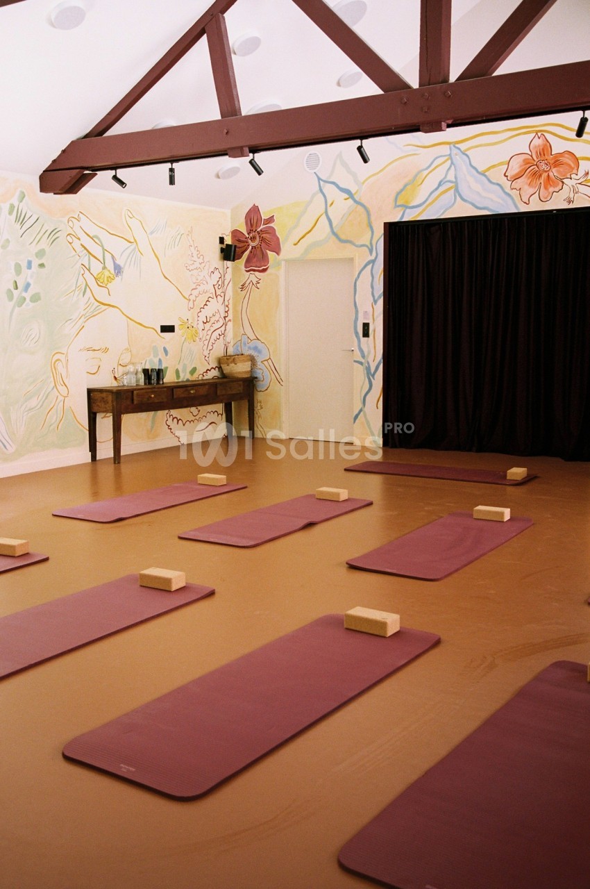Salle de yoga avec tapis violets alignés, briques en bois et mur décoré de peintures florales et abstraites.