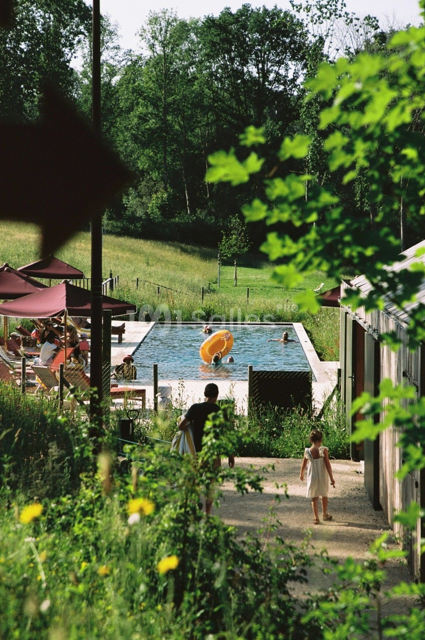 Vue d'une piscine extérieure entourée de verdure, avec des personnes se relaxant et des enfants jouant.