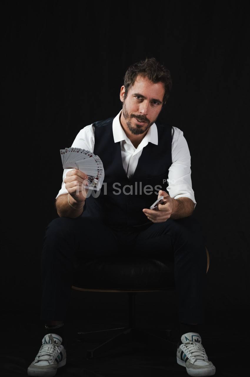Un homme assis en costume décontracté, tenant un éventail de cartes dans une main, sur fond noir.