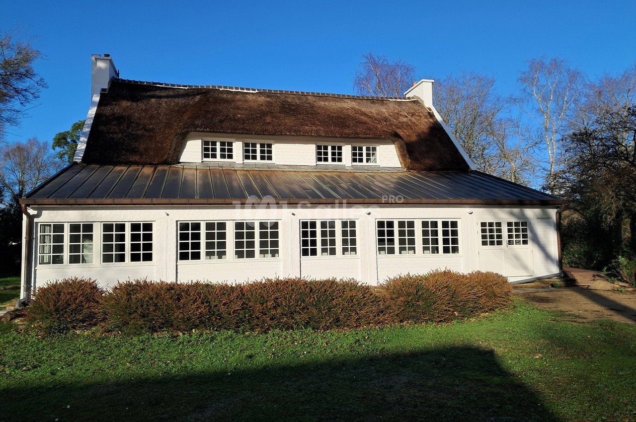 Maison blanche avec toit de chaume, grandes fenêtres et jardin, éclairée par le soleil sous un ciel bleu.