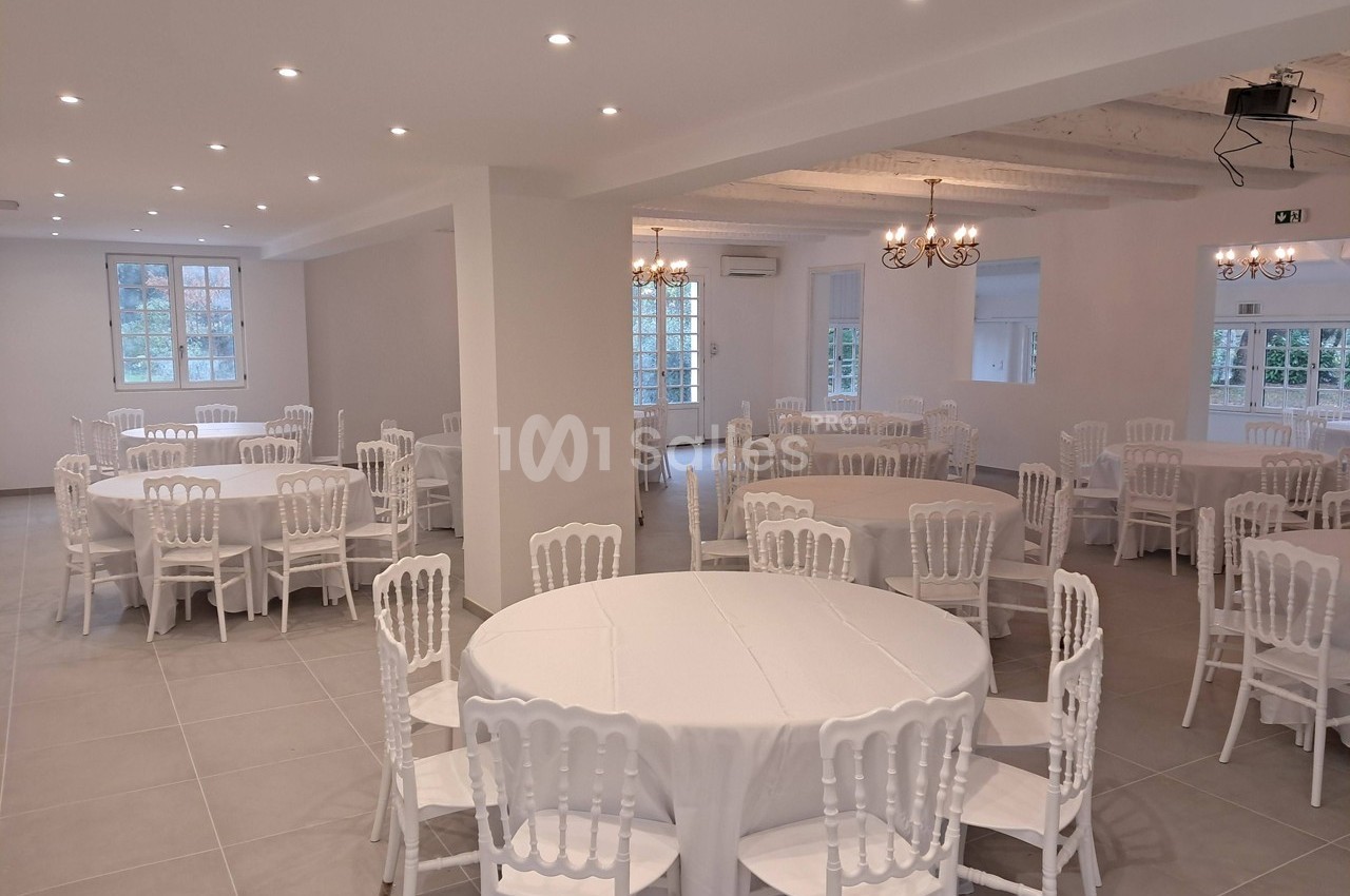 Salle lumineuse avec tables rondes dressées de nappes blanches et chaises blanches, éclairée par des lustres et spots.
