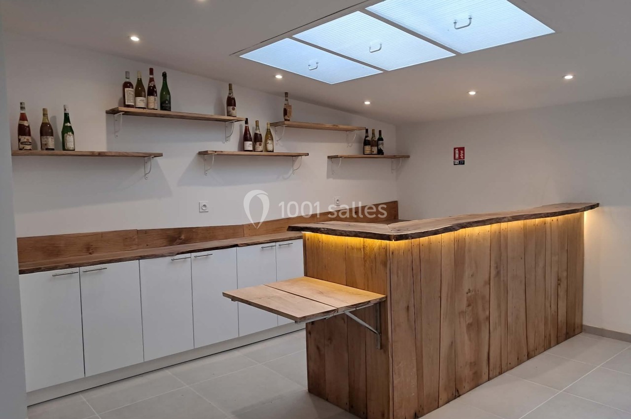 Bar en bois avec comptoir éclairé, étagères murales garnies de bouteilles et plafond avec puits de lumière.