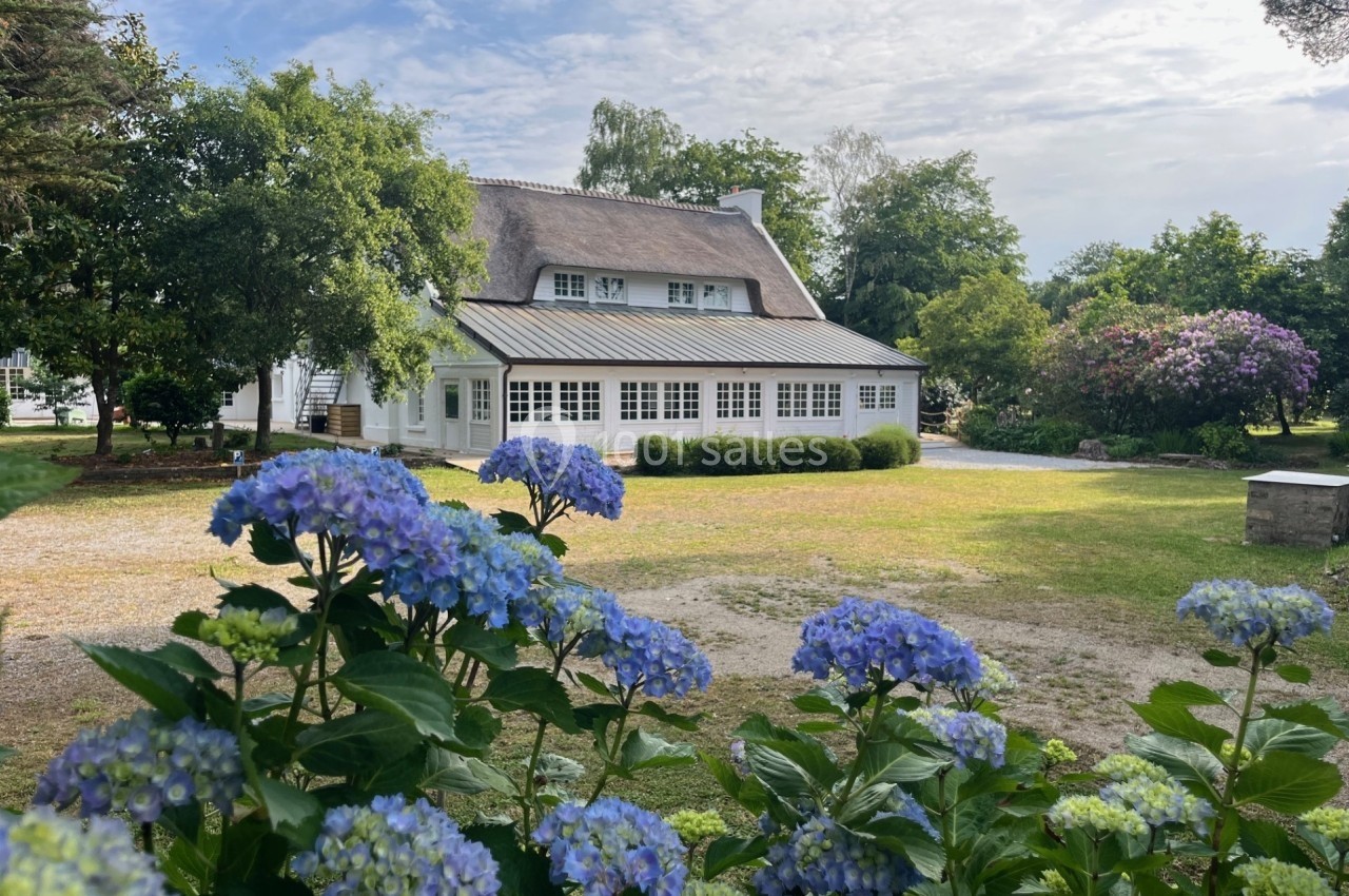 Maison blanche au toit de chaume entourée de verdure, avec des hortensias bleus au premier plan.