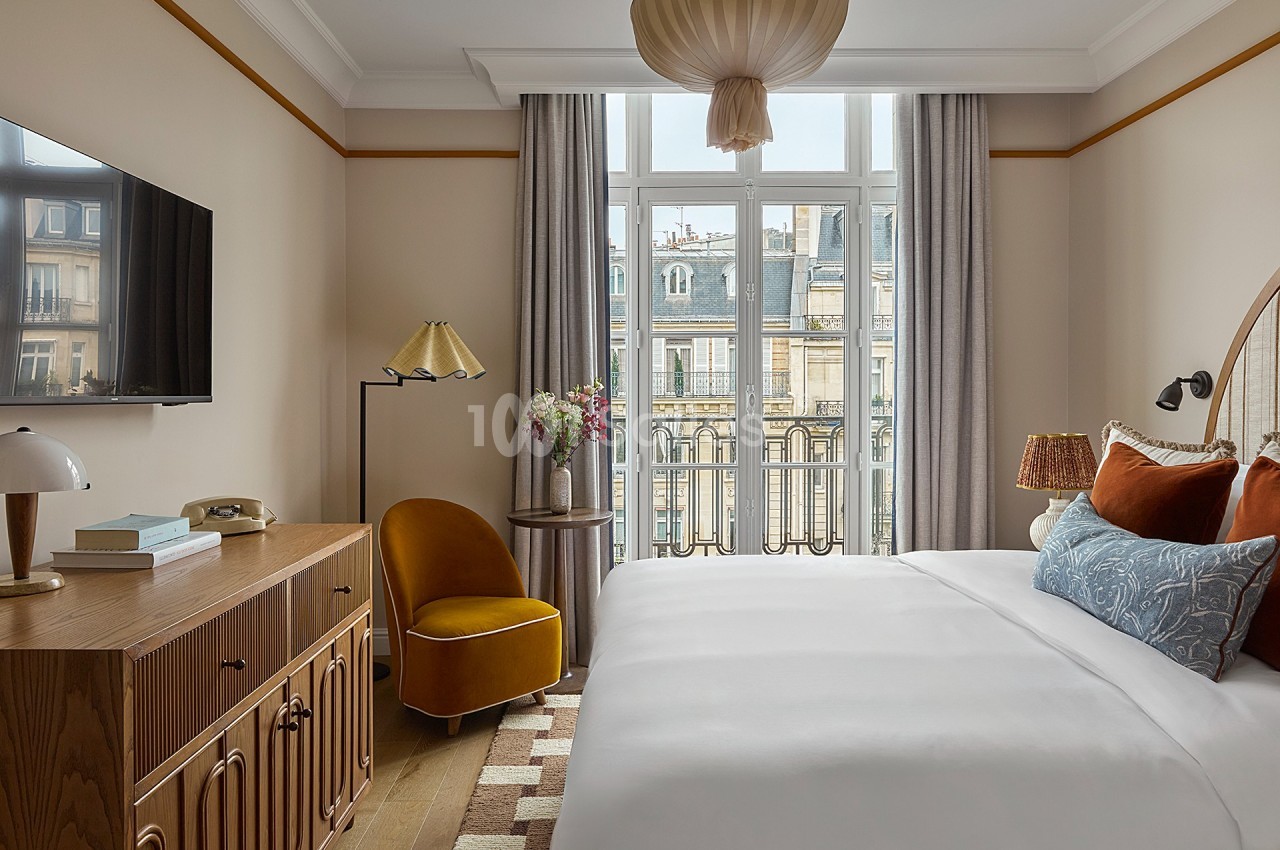 Chambre lumineuse avec lit double, fauteuil jaune, commode en bois et grande fenêtre donnant sur des immeubles parisiens.