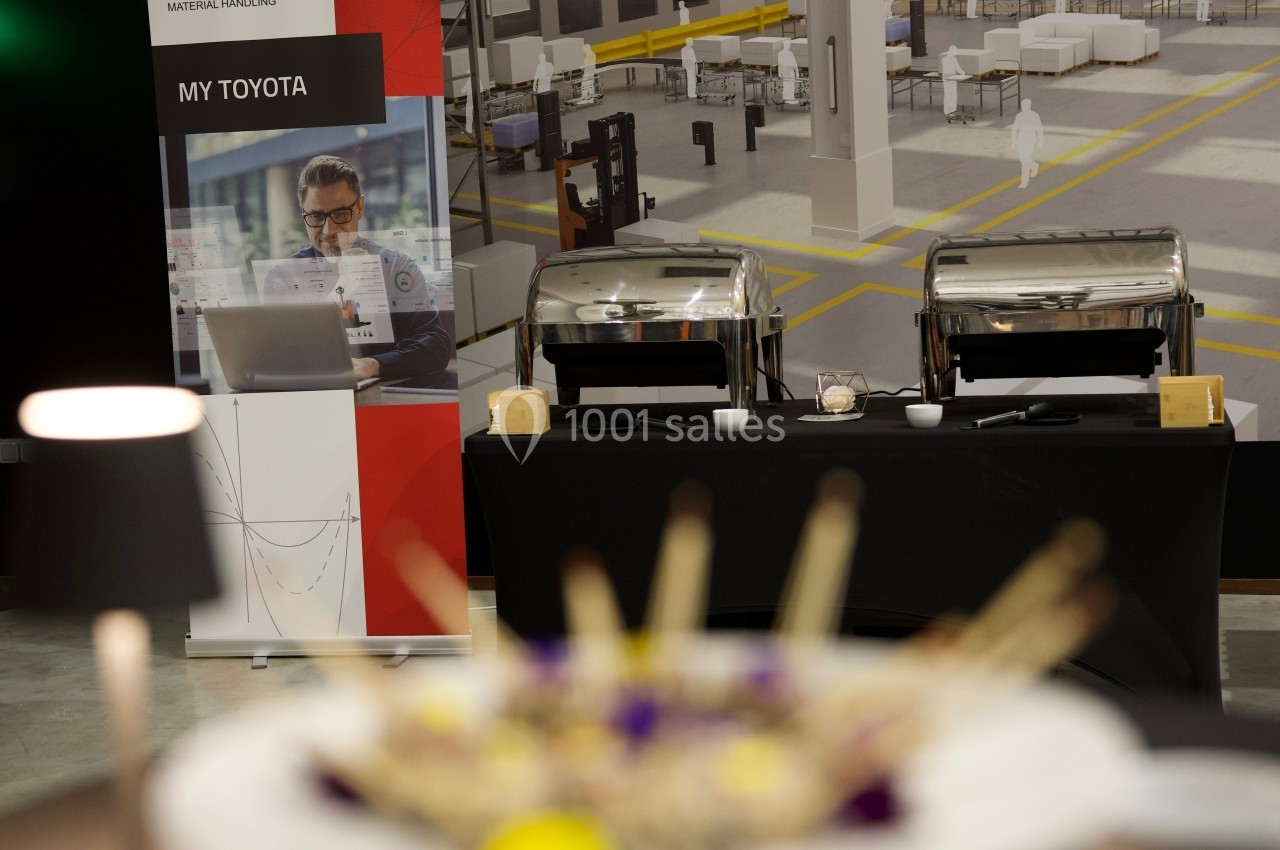 Buffet avec plats sous cloches en inox, devant une bannière Toyota et un fond d'entrepôt industriel.