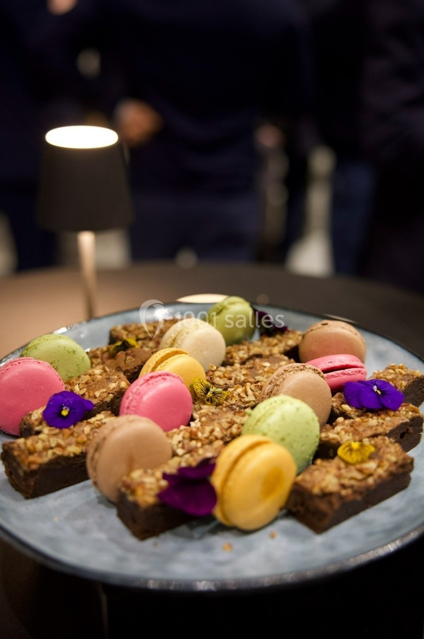Assortiment de macarons colorés et brownies garnis de fleurs comestibles, présenté sur une assiette bleue.